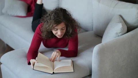 Young woman with red pullover reading hardcover book on grey couch at home. Stock Footage 142843358