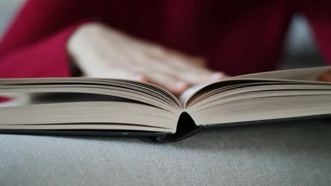 Young woman with red pullover reading hardcover book on grey couch at home. Stock Footage 142843559