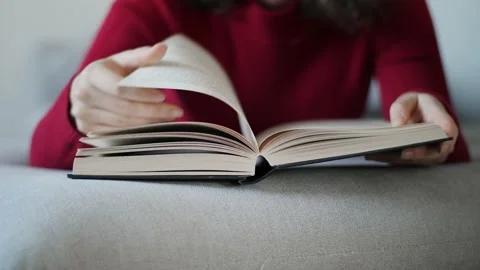 Young woman with red pullover reading hardcover book on grey couch at home. Stock Footage 142843699