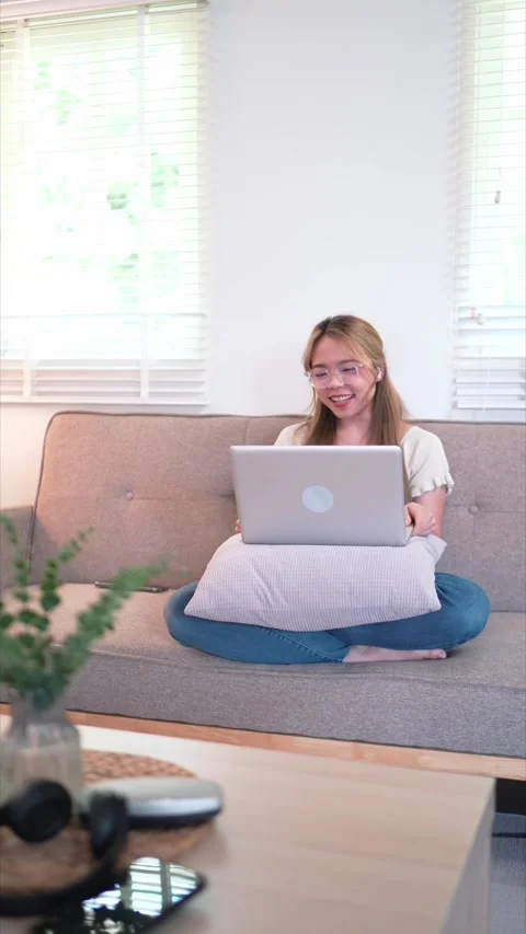 A young woman sits on a couch, using a laptop. She looks engaged in her wor.. Stock Footage 288732175