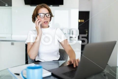 Young woman sits at the kitchen table using a laptop and talking on a cell ph Stock Photos