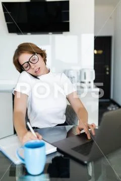 Young woman sits at the kitchen table using a laptop and talking on a cell ph Stock Photos
