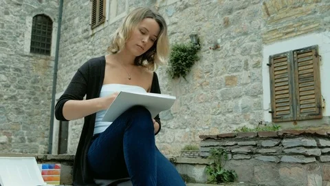 Young woman sits on stones of old city and paints with watercolors. Attractive Stock Footage