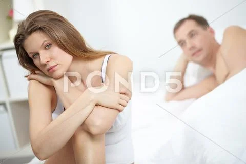 A young woman sitting on bed upset with her lying husband in the background Stock Photos