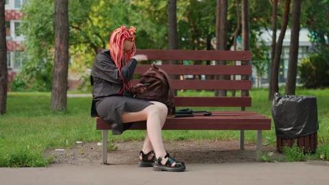 Young woman sitting on bench checking backpack, pink dreadlocks, urban park 库存影片 330717584