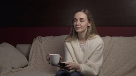 Young woman sitting on couch drinking tea, switching TV channels on remote Stock Footage 126571953