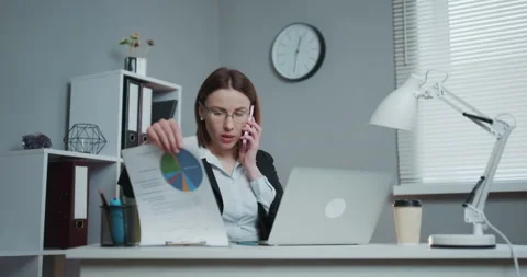 Young woman sitting at desk using laptop, answering call, talking on the phone Stock Footage 194486053