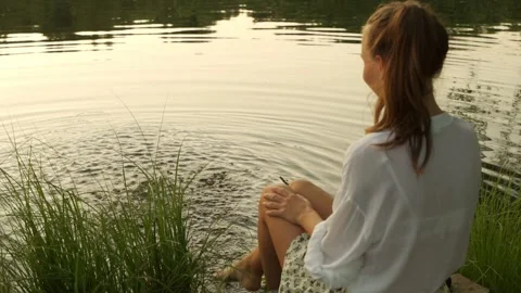 Young woman sitting on dock splashing bare legs in lake. Women's feet in the Stock Footage