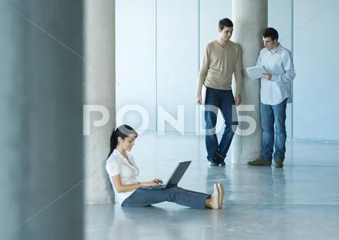 Young Woman Sitting On Floor Using Laptop, Two Men Standing Takling About