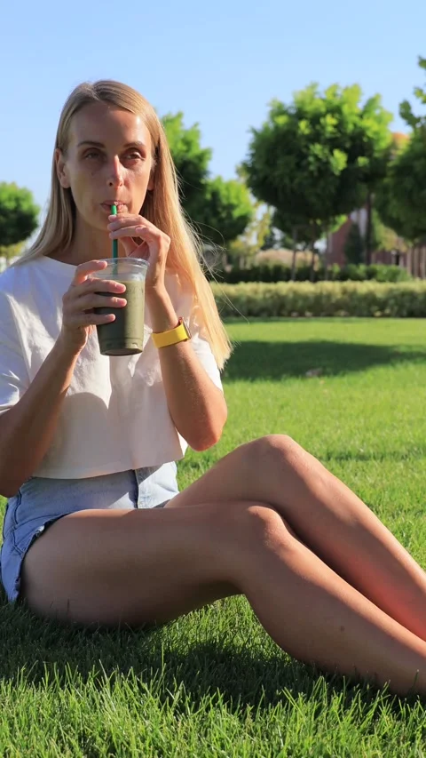 Young woman sitting on grass drinking iced matcha latte with straw on sunny day Stock Footage 315744248