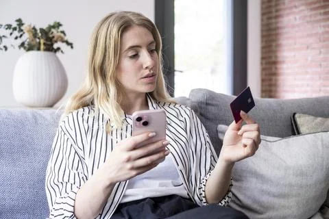 Young woman sitting on a sofa using a smartphone and holding a credit card .. Fotos Stock