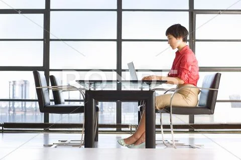 Young Woman Sitting At Table By Window, Using Laptop, Side View