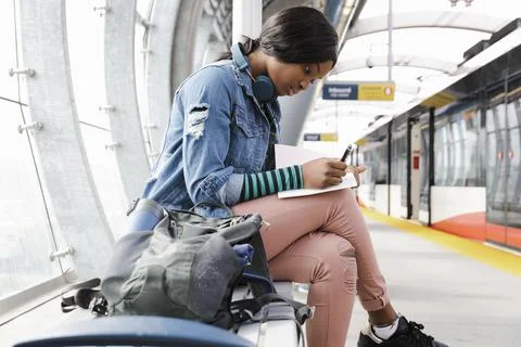 Young woman sitting on train platform making notes in book Stock Photos