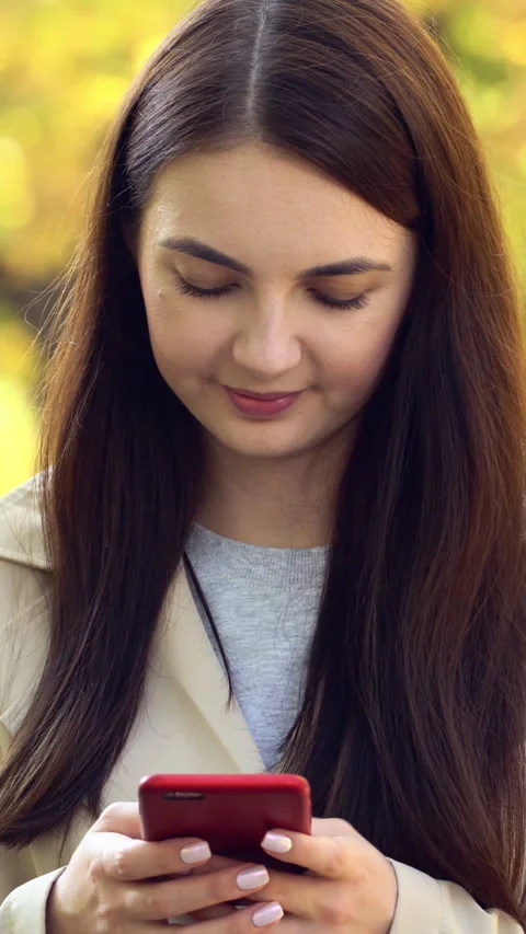 A young woman smiles while checking her red smartphone outdoors in autumn with Video stock 347483590