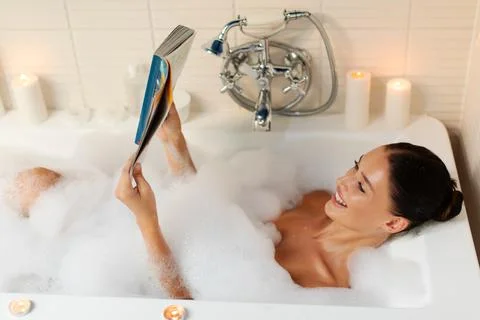 Young woman smiling while reading magazine in foamy bathtub at home Stock Photos
