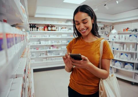 Young woman smiling while using smartphone standing against shelf in pharmacy Stock Photos