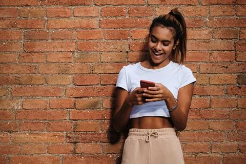 Young woman smiling while using a mobile phone standing outdoors against a brick Stock Photos