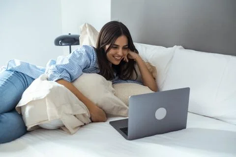Young woman smiling while using laptop on bed at home, relaxed remote work .. 스톡 사진