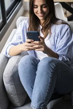 Young woman smiling while using smartphone on couch at home in casual outfit Foto stock