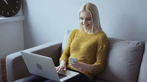 Young woman on sofa using her smartphone and laptop for online banking Stock Footage 64333588
