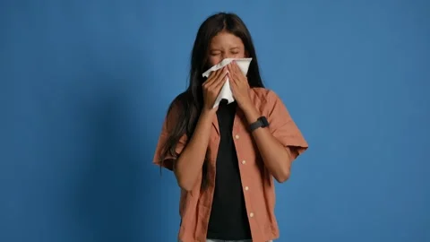 Young woman standing against blue background sneezing due to cold, wiping her Stock Footage 337972353