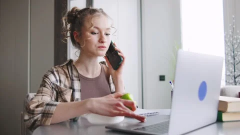 Young woman talks on phone while working on laptop and eating apple in home Stock Footage 346185244