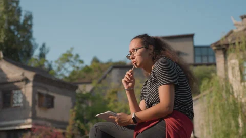 Young woman tasting snack while jotting notes, playful expression along canal Stockbeeldmateriaal 331469337