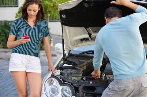 Young woman text messaging while her husband looking into car engine Stock Photos