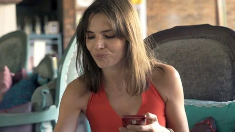 Young woman texting on smartphone and drinking beverage on sofa in trendy cafe Stock Footage