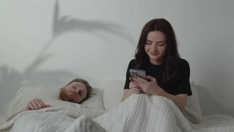 Young woman texting while her husband is sleeping. Beautiful girl typing Stock Footage 233967927