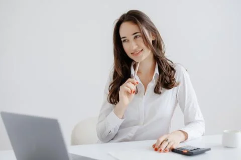 Young woman thinking while working at desk with laptop, calculator, and coffee Stock Photos