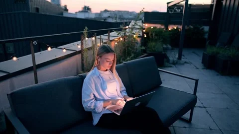 Young woman typing on laptop while sitting on a rooftop terrace at dusk. Cozy Stock Footage 320904045