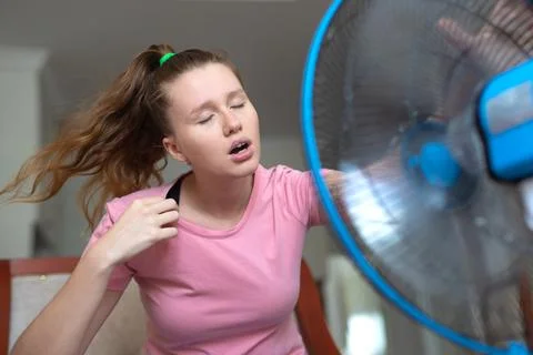 Young woman using electric fan at home in living room, sitting on couch cooling Stock Photos