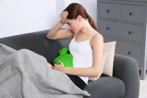 Young woman using hot water bottle to relieve cystitis pain on sofa at home Stock Photos