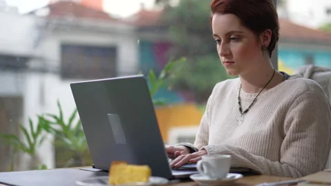 Young woman using laptop computer seated at coffee shop by window. 20s red ha Stock Footage 205844863