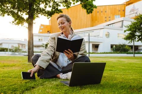 Young woman using note book and laptop while sitting on grass outdoors Stock Photos