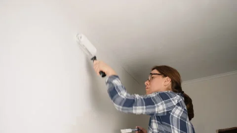 Young woman using a roller to paint the top edge of a wall near the ceiling. She Stock Footage 312649183