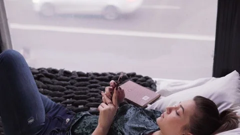 Young woman using smartphone on bed by window overlooking city street Stock Footage 86086361