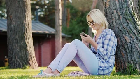 A young woman is using a smartphone. Sits on the grass under a tree in the Stock Footage 80018705