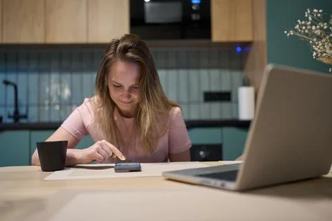 Young woman using smartphone at table with laptop in modern kitchen Stock Photos
