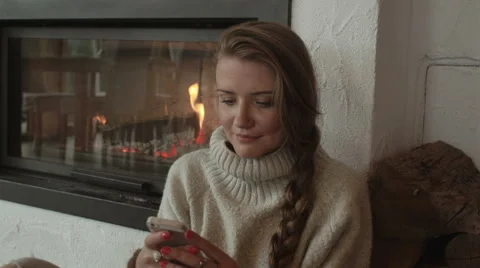 Young woman using smartphone while sitting near fireplace at home. 動画素材 56318662