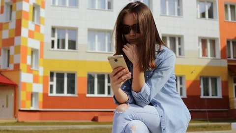 Young woman using smartphone while sitting on parapet near business building 스톡 동영상 128571758