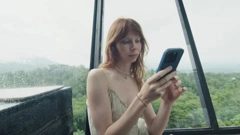 Young woman using smartphone while relaxing in modern house during rain Stock Footage 312999681