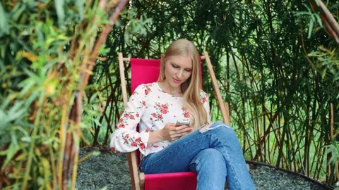Young woman using smartphone while seated in garden with bamboo foliage Video stock 340565307
