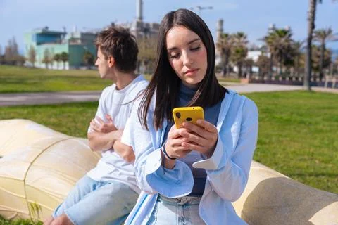 Young woman using smartphone while boyfriend looks away in sunny park Stock Photos