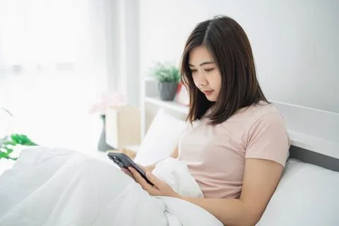 Young woman using smartphone while sitting in bed, cozy atmosphere with sof.. Stock Photos