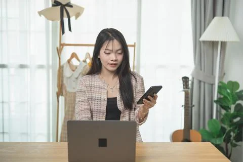 Young woman using smartphone while sitting at desk with laptop, clothes rac.. Fotos de archivo