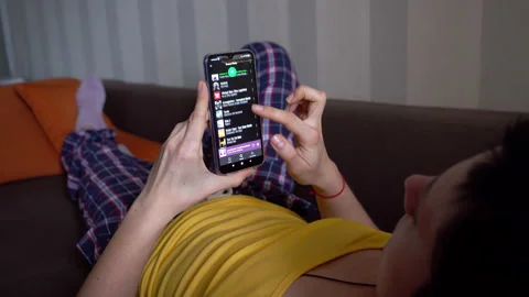 Young woman using Spotify app on smartphone at home, lying on a couch.  Stock Footage 145761910