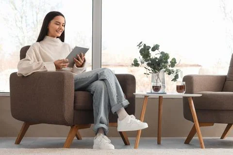 Young woman using tablet computer in armchair near large window at home Stock Photos