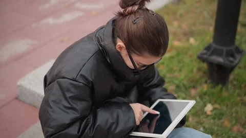 Young woman using tablet outdoors on city sidewalk with grass background with Stock Footage 346729503